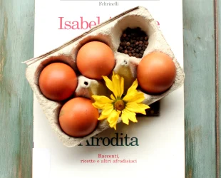 Libro Afrodita di Isabel Allende sul quale è poggiata una scatola di cartone con quattro uova; nei buchi senza uova ci sono pepe in grani e una gerbera gialla. Il cartone è poggiato in modo da coprire l'immagine, ma si intravedono il titolo e il nome dell'autrice.