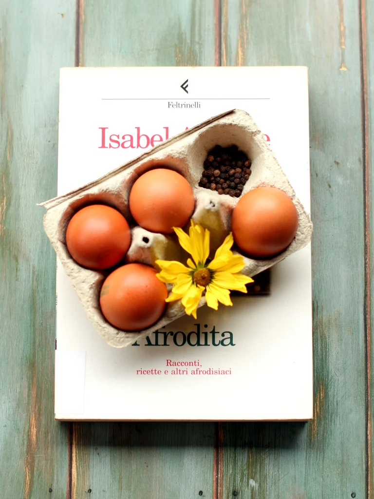 Libro Afrodita di Isabel Allende sul quale è poggiata una scatola di cartone con quattro uova; nei buchi senza uova ci sono pepe in grani e una gerbera gialla. Il cartone è poggiato in modo da coprire l'immagine, ma si intravedono il titolo e il nome dell'autrice.