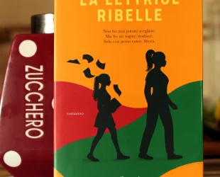 Copertina del libro La lettrice ribelle di Ishi Robinson: sfondo arancione con due strade stilizzate, una strada verde e una rossa poco dietro; le sagome nere di una donna e una ragazza camminano verso destra. La copertina è poggiata a un contenitore bordeaux a pois bianchi, lascia visibile la scritta Zucchero sulla sinistra, ruotata in verticale verso destra.