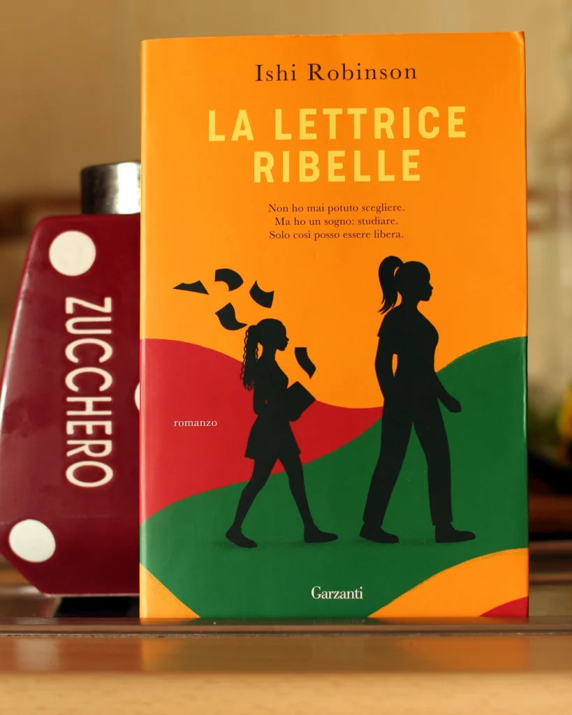Copertina del libro La lettrice ribelle di Ishi Robinson: sfondo arancione con due strade stilizzate, una strada verde e una rossa poco dietro; le sagome nere di una donna e una ragazza camminano verso destra. La copertina è poggiata a un contenitore bordeaux a pois bianchi, lascia visibile la scritta Zucchero sulla sinistra, ruotata in verticale verso destra.