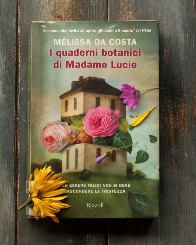 Copertina del libro I quaderni botanici di Madame Lucie, di Melissa Da Costa. C'è il disegno di una casa in collina attraversata, tra il primo e il secondo piano, da fiori più grandi delle finestre; alcune foglie svolazzano intorno. Ho decorato con una gerbera gialla e un fiore di malva. Questo è il libro che ho scelto per parlare di scrittura intima.